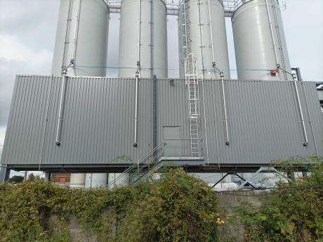 Quatre silos rigides industriels blancs alignés sur une plateforme métallique avec échelle de sécurité et tuyauterie, devant une façade à panneaux gris.