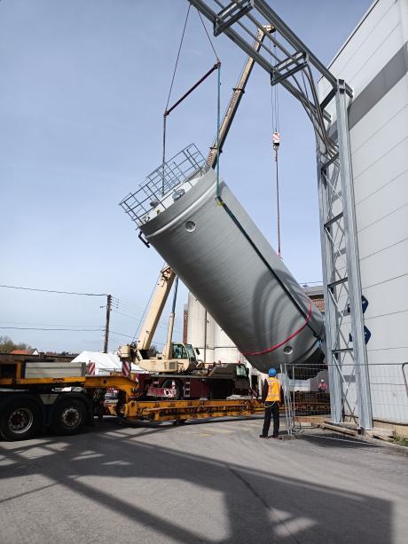 Ouvrier en gilet haute visibilité guidant l'installation d'un silo rigide industriel incliné par une grue mobile montée sur un camion plateau sur site de production.