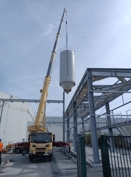 Grue mobile en train de positionner un silo rigide industriel blanc sur une charpente métallique extérieure sur site de production.