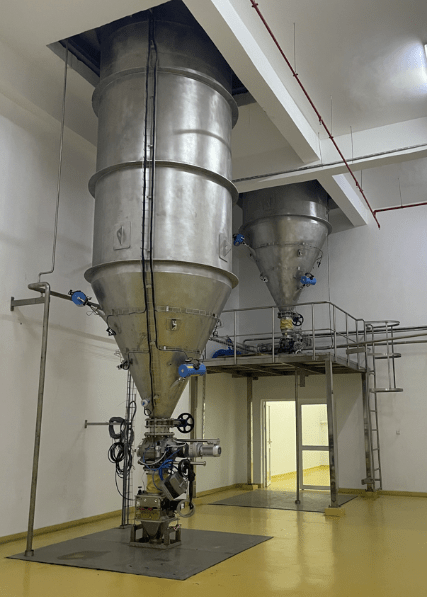 Deux silos rigides en acier inoxydable installés en intérieur sur une plateforme métallique avec vannes et tuyauterie pour l’alimentation et la régulation d’un process industriel.