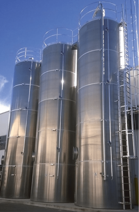 Trois silos rigides industriels en acier inoxydable brillants, équipés d’échelles et de garde-corps, installés à l’extérieur sous un ciel bleu.